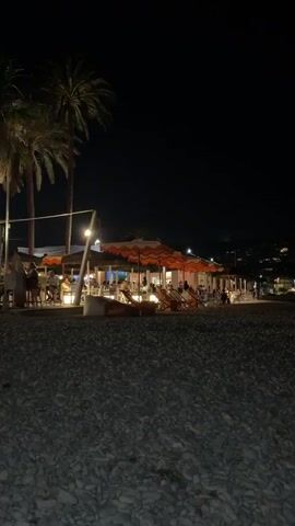 The video shows a nighttime scene on a pebble beach. In the background, a brightly lit restaurant or beach club is visible, with people seated at tables under orange and white striped umbrellas. Palm trees are also present. The foreground is dominated by the dark, textured surface of the beach. The overall impression is of a lively evening at a seaside establishment.