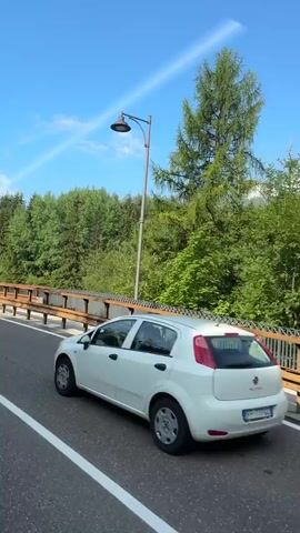 The video shows a roadside view from a moving vehicle on a sunny day. A grey camper van passes by on the road in the foreground. The road has white lines marking the lanes. To the right of the road, there is a metal guardrail with a wooden top, separating the road from a densely wooded area with tall green trees and some mountains in the distance. A streetlight is visible on the left side of the road. The sky is blue with some white clouds, and a faint white line, possibly a contrail, is visible across the sky. The scene appears to be a scenic route, possibly in a mountainous region.