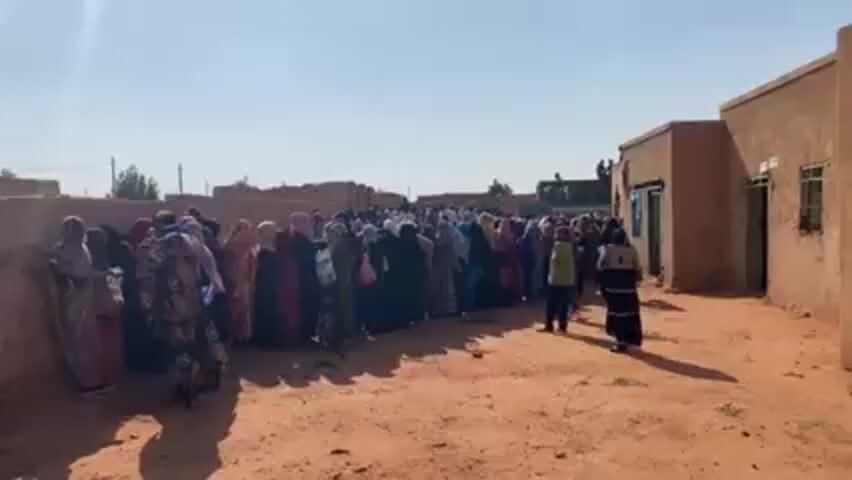 A long line of people is standing outside a small building, likely waiting to enter. The majority of the people are women, many wearing traditional clothing, including headscarves. The line stretches into the distance, suggesting a large number of people are waiting. The setting appears to be a rural or village area, characterized by sandy ground and simple, mud-brick buildings. The sky is clear and bright. There are a few children also present in the crowd.