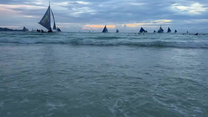 The video shows a beach scene with the ocean in the foreground and sailboats in the background. The water is a clear blue-green and there are gentle waves breaking on the shore. Several sailboats with triangular sails are scattered across the ocean, some with people visible on them. The sky is cloudy with hints of orange and pink from a sunset. The overall atmosphere is peaceful and serene.