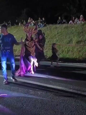 A nighttime outdoor parade is depicted. Several people are visible in the foreground, including a man in a blue t-shirt and jeans, two individuals in elaborate costumes with headpieces and long flowing garments, and a child. The costumed individuals appear to be part of a performance, possibly a parade or festival. In the background, a crowd of spectators is seated on a grassy embankment, watching the procession. The environment appears to be a road or street, with a grassy hill and trees behind the spectators. The lighting is dim, suggesting it is dark outside, and there are some artificial lights illuminating the parade participants.