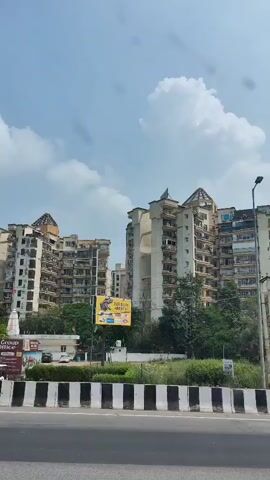 The video shows a view from a vehicle driving past a row of tall apartment buildings. The buildings are multi-story with many balconies. Some of the buildings have visible signage. There are trees and greenery in front of the buildings, and a highway barrier with black and white stripes runs along the road. The sky is cloudy with some patches of blue. In the foreground, parts of another vehicle, including its windshield and side mirror, are visible. There is also a street lamp on the right side of the frame. A billboard with an advertisement and a small temple-like structure with a flag are also visible between the buildings and the road.