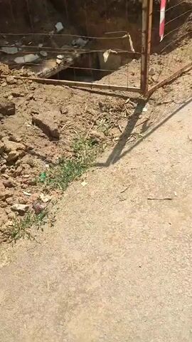 The video shows an outdoor scene with a section of exposed pipes in a dirt trench. The trench is partially surrounded by a wire mesh fence, with some red and white caution tape tied to a rusty metal post. The pipes appear to be old and weathered, with some dirt and debris on them. The surrounding ground is made of dry soil and rocks. There are no people visible in the video.