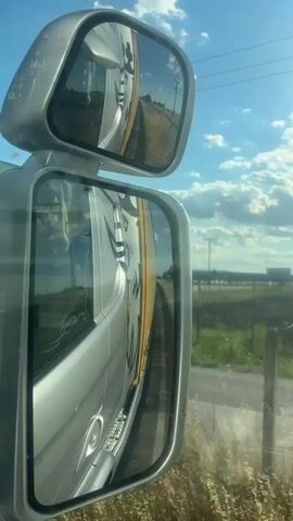 This media file presents a view through the side mirrors of a vehicle, likely a truck or bus, as it travels along a road. The primary subjects are the vehicle itself, reflected in the mirrors, and the passing scenery.

The larger, lower mirror shows a significant portion of the vehicle's side, which appears to be silver with yellow accents and black lettering. The lettering is stylized and appears to be part of a logo or company name, though it is difficult to read clearly. The smaller, upper mirror reflects the road ahead and a curved horizon, suggesting movement.

The setting is an outdoor, rural environment. In the background, visible beyond the vehicle and through the mirrors, are fields of green grass and crops, with some dry vegetation closer to the road. There are power lines stretching across the sky and several utility poles. The sky is a bright blue with scattered white clouds, indicative of daytime. The lighting suggests it is a sunny day, possibly late morning or afternoon.

The activity taking place is travel. The perspective from the side mirrors captures the continuous motion of the vehicle and the shifting landscape. There are no people visible in the immediate scene, only reflections and the vehicle's interior reflected in the main mirror.

The overall mood is one of movement and observation, with the mirrors acting as a frame for the unfolding journey. The location context of Orestiada, Greece, places this scene in a specific geographical area, suggesting a journey through the Greek countryside. No overt emotions are conveyed, but the clear weather and open scenery might evoke feelings of freedom or progression. There are no legible signs or text other than the branding on the vehicle itself.