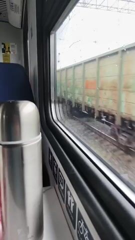 The video shows a view from inside a train car. In the foreground, to the left, a stainless steel thermos is visible. The window of the train looks out onto a railway track where a freight train is passing by. The freight train consists of several boxcars, some are green and some are brown. The movement of the passing train causes a blurring effect in the video. The window also has a strip of stickers along the bottom edge, depicting various icons such as a television, a person in a wheelchair, and a bicycle. The lighting appears to be overcast or daytime with diffused light. The overall impression is a snapshot of a journey on a train, observing the passing scenery.