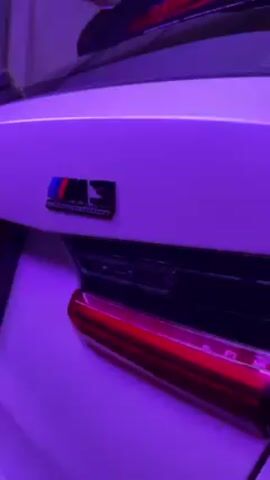 The video shows the rear of a white BMW M3 Competition, illuminated by purple lighting. The M3 Competition badge is visible on the trunk lid.  The car's taillights are illuminated with red LEDs. A small portion of a spoiler is visible above the trunk.