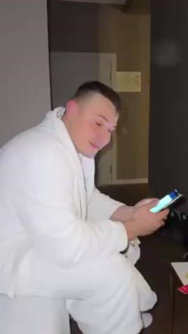A man wearing a white bathrobe is sitting on a bed in a room. He is looking at his phone. He turns and looks at the camera, smiling. He holds up his phone, displaying the screen to the camera. The screen shows a picture of a person wearing a white bathrobe and sleeping on a white bed. The background is a room with dark walls and a white door.