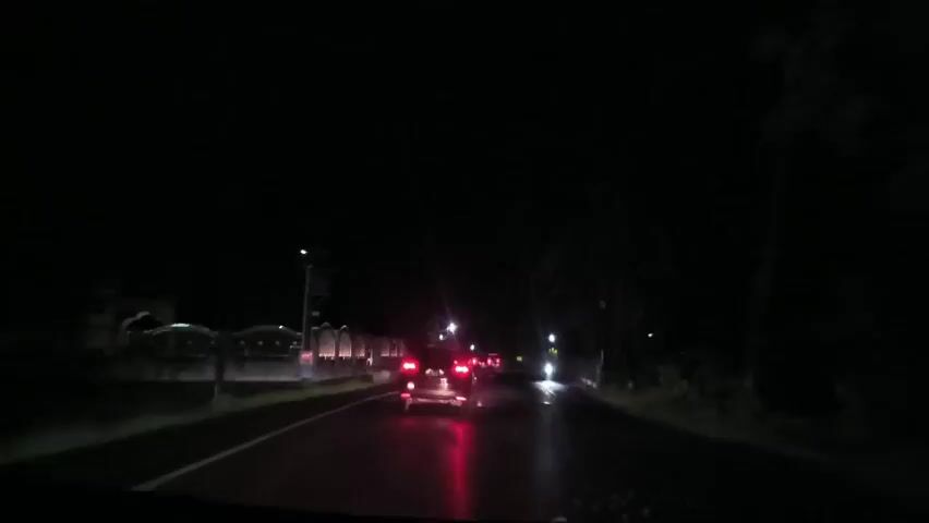 The video shows a car driving on a dark road at night, following another car with illuminated taillights. Streetlights are visible on both sides of the road. A wall or fence runs along the left side of the road, and vegetation can be seen on both sides. The road appears wet, reflecting the car's taillights.