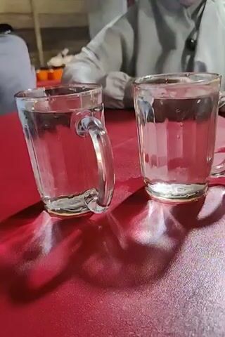 The video shows the interior of a simple restaurant, focusing on two glasses of water on a red table, and a young boy sitting at the table.  The glasses are mostly full. The boy is wearing a light grey shirt. He looks up at the camera with a blank expression.  Other people are sitting at tables in the background, visible through a window.  The restaurant seems to be constructed of wood.  The lighting is somewhat dim, creating strong shadows from the glasses.