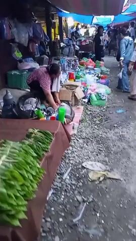 A bustling outdoor market is depicted, with numerous people browsing and shopping under colorful awnings. The ground is covered in gravel and debris, suggesting a rural or informal setting. In the foreground, a vendor is arranging a display of fresh green leaves, likely produce, on a stall. To the right, a woman in a yellow shirt and blue jeans walks past, seemingly a shopper. The market appears to be well-stocked with various goods, including plastic containers, kitchenware, and bags. The overall atmosphere is lively and active, indicative of a busy marketplace.