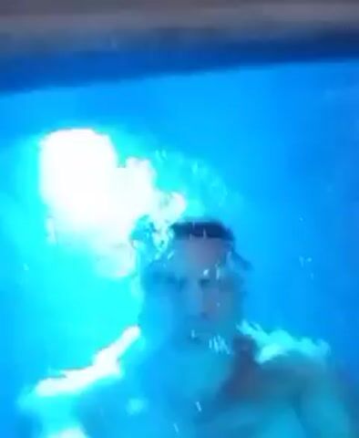 A man emerges from underwater in a swimming pool. The pool is lit with a bright, blue light, creating a surreal ambiance.  He is shirtless and appears to be looking directly at the camera.  The background shows a white wall and some trees, suggesting it is nighttime.  The man seems to be treading water.