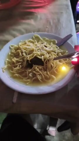 A close-up video shows a bowl of noodles with broth. A hand holding chopsticks is lifting a portion of the noodles from the bowl. In the background, a table is visible with several people seated around it. There are bottles and other food items on the table. The lighting suggests an outdoor or dimly lit setting.