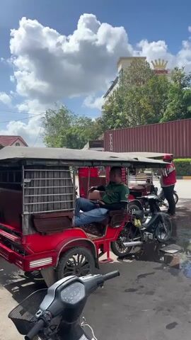 The video shows a street scene in Cambodia with tuk-tuks and motorcycles parked near a puddle. One man is sitting in a tuk-tuk, another is standing near his motorbike and speaking on the phone, and a third man is walking across the street. The scene includes a view of the Crown Royal Casino in the background, along with several buildings and shipping containers. The sky is partly cloudy.