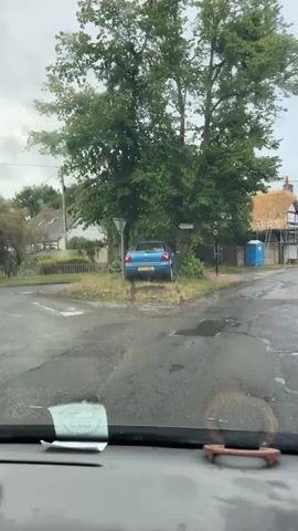 The video shows a blue Volkswagen Polo car parked off a paved road, partially obscured by trees and bushes. The car's front is facing a large tree, and its rear is visible to the viewer. The license plate reads "WD04 MMF". The camera is inside another vehicle, looking out through the front windshield, which has some raindrops on it. Puddles are visible on the road surface. In the background, there are houses and what appears to be a construction site with a blue portable toilet. A wooden signpost points to "LONG ROW" and "HADDON", indicating a rural or village setting.