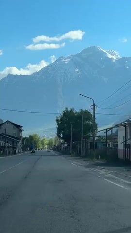 The video shows a road in a mountainous region with a clear blue sky and scattered clouds. A large, snow-capped mountain dominates the background. On the left side of the road, there are buildings, one of which is a multi-story structure. On the right side, there are more buildings, fences, and utility poles with wires. A few cars are visible on the road, moving away from the camera. A tree with green foliage is situated in the middle ground, to the right of the road. The road itself is paved and appears to be in fair condition with some cracks. The overall scene suggests a scenic, possibly rural or semi-urban, environment with a prominent natural landscape.