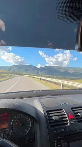 The video is captured from the perspective of the driver inside a car. The driver's hands are visible on the steering wheel as the car is in motion on a paved road. The road curves to the left, with a guardrail lining the right side. In the distance, rolling hills and mountains are visible under a bright blue sky with scattered white clouds. The car's dashboard is also in view, showing the speedometer indicating a speed of approximately 100 km/h and other information displayed on a digital screen. The sun is shining, casting light on the landscape. The overall impression is a scenic drive through a rural or mountainous area.