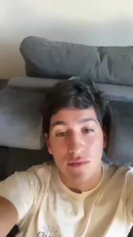 A young man is lying on a gray couch, looking directly at the camera. He has dark hair and light skin. He is wearing a light-colored t-shirt with a graphic on it. He appears to be startled, as he opens his mouth wide and makes a noise. The background is a plain, light-colored wall.