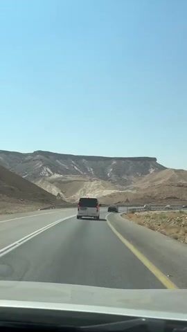 The video shows a view from inside a vehicle traveling on a paved road through a desert landscape. A white minivan is visible ahead, driving in the same direction. Two other vehicles are also on the road, one behind the minivan and another further ahead. The road has a yellow line marking the edge and white lines dividing lanes. On either side of the road, there are guardrails and arid, hilly terrain with sparse vegetation. In the distance, prominent flat-topped mountains rise against a clear blue sky. Several warning signs are visible along the roadside. The interior of the vehicle, including the dashboard and windshield wipers, can be seen in the foreground, indicating a first-person perspective. A child's voice can be heard in the background, calling out "Papa."