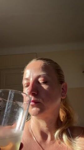 A close-up, low-angle shot shows a woman with blonde hair pulled back in a ponytail, drinking from a clear glass. The sun, casting long shadows, illuminates her face from the left, highlighting the texture of her skin and her closed eyes as she sips. She is wearing a white tank top and a delicate necklace. The glass has visible bubbles and a light-colored liquid. Text is visible on the glass: "MONKEY SHOULDER". The background appears to be an indoor setting with a ceiling and a wall, suggesting it is daytime, possibly late afternoon due to the lighting. The overall mood is serene and relaxed. The location context is Dobratycze, Poland.