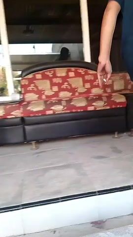 The video shows a person from behind, standing on a tiled platform. They are wearing a dark t-shirt and patterned shorts. The person is barefoot and holding a cigarette in their left hand and a small yellow object in their right hand. They appear to be leaning slightly forward. The environment is an indoor/outdoor space, with a sofa visible to the left and a doorway or opening to the right. The flooring is tiled, with some red and white discoloration on the edges.