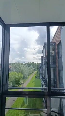 The video shows a view from a balcony or enclosed porch looking out at a street lined with trees and apartment buildings. The sky is cloudy. The video is handheld and shows a slight wobble.