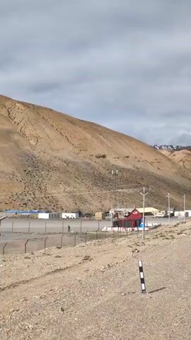 This is a landscape video. The video shows a desolate mountainous terrain with a cloudy sky. In the foreground, a dirt road is visible. Further back, there are some buildings with red roofs, possibly a small complex or settlement. There are also fences, utility poles, and traffic signs. The overall environment appears dry and barren, characteristic of a high-altitude desert or steppe region. There are no visible people or animals in the frame, and no discernible actions or interactions are taking place.