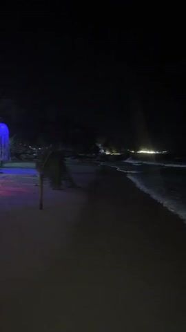 The video shows a dark beach scene at night, panning from right to left. On the right, dark ocean waves are visible with distant lights along the horizon. As the camera pans left, a section of the sandy beach is revealed, illuminated by numerous colorful, jellyfish-shaped lanterns hanging from unseen structures or trees. The lanterns are red, blue, green, pink, and purple. Below the lanterns, several people are seen sitting on low cushions or beanbags around small tables or ground-level lights. One person stands near a bright light source on the ground. More light sources and structures are visible in the background, further down the beach.