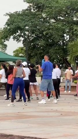 A group of people are exercising outdoors. A woman in the foreground is wearing a grey tank top and blue leggings. To her left, a woman in a white t-shirt and dark pants is partially visible. Behind them, a group of people are dancing in a line, facing away from the camera. A man in a blue t-shirt and denim shorts is in the center of the group, leading the dance. Other participants are wearing t-shirts and athletic pants. The scene takes place in a park with trees and a paved area.
