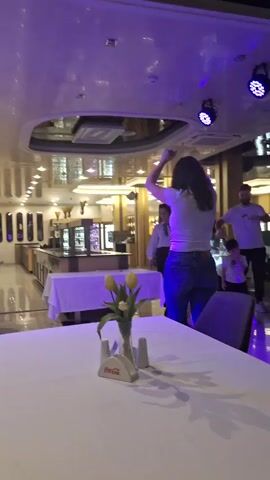 The video shows a group of people dancing in a restaurant, including a woman with dark curly hair, wearing jeans and a white top, and a woman with blond hair, wearing a black floral shirt and black pants. A man with a beard and a little boy are also present, along with other people who are out of focus in the background. The scene is illuminated by colored lights that change between red, green, and blue. There are tables with white tablecloths and centerpieces with flowers in the foreground. The restaurant has an open layout with buffet-style food stations in the background.