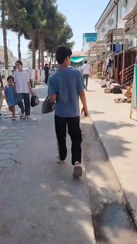 A young boy with dark hair wears a blue t-shirt and black pants with a white stripe. He carries a black and white striped bag in his right hand as he walks down a sidewalk. Behind him, a girl in a blue dress and a woman in a white t-shirt and patterned pants walk together. The girl is holding a black bag. An older man in a light-colored shirt and pants is walking in front of them. Trees line the left side of the sidewalk, and buildings are visible on the right. Other people are walking in the distance. The ground is paved with light-colored stones in a circular pattern. Sunlight casts shadows on the sidewalk.