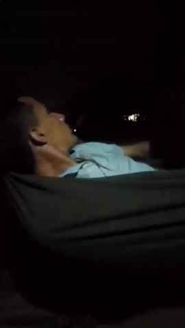 A man is lying in a hammock at night. He is wearing a light blue collared shirt and has short dark hair. He looks at his phone and then looks around. A light is visible in the distance behind him.