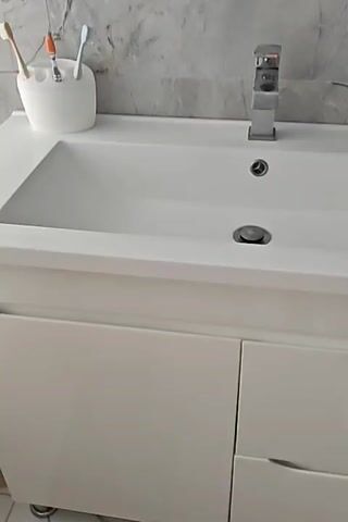 The media file shows a close-up, panning view of a modern white bathroom vanity. The sink is pristine white ceramic with a chrome faucet. To the left of the sink, a white holder contains toothbrushes, some with colorful bristles. To the right of the sink, there is a small white soap dish holding a bar of soap. The vanity itself is white with a glossy finish, featuring two drawers with integrated handles. The wall behind the vanity is tiled with a grey marble pattern. The overall scene depicts a clean and well-maintained bathroom. There is no visible text in the media file. The lighting suggests it is daytime, and the environment appears to be indoors. There are no people visible, and no specific event is taking place beyond the static presentation of the bathroom fixture. The location context of Izmail, Ukraine, is noted but not visually evident in the media itself.