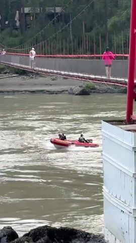 A red inflatable boat with two people is seen on a fast-moving river. The boat is heading towards the right of the frame, creating a wake. In the background, a suspension bridge with red railings is visible, spanning across the river. Several people are walking on the bridge. The riverbanks are lined with trees and vegetation. The water is murky brown, indicating a possibly strong current or sediment.