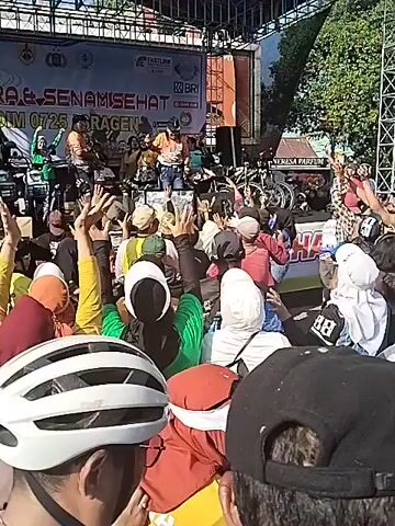 A large crowd of people, mostly in cycling gear, are gathered in front of an outdoor stage under a clear sky. Many are raising their arms in the air. On the stage, several individuals are performing or leading an activity. Banners with text and logos, including "GOWES GEMBIRA & SENAM SEHAT" and "KODIM 0725 SRAGEN," are visible behind them. Some bicycles are also seen on the stage. The event appears to be a lively gathering or festival.