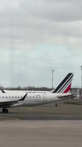 The video shows two Air France airplanes on a tarmac against a cloudy sky. The airplanes are primarily white with blue, white, and red accents on their tails. The aircraft closer to the camera has the registration number F-HBCQ visible on its body. A small control tower and some red and white striped poles are visible in the background. Another plane is distantly visible in the sky. The view is framed as if taken through a window.