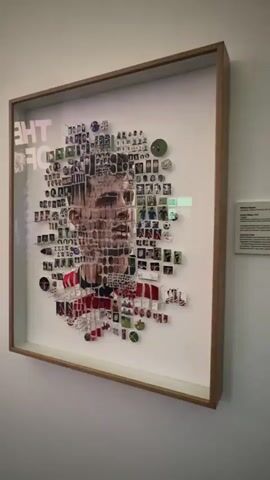 A framed artwork is displayed on a white wall. The artwork is a mosaic portrait of a person's face, composed of many small photographs and other small items. Some of the smaller items appear to be medals or badges. The portrait is large and detailed, with the smaller pieces arranged to form the contours of the face and hair. In the background of the artwork, text is visible, which appears to read "THE OF." To the right of the framed artwork, a small white plaque with black text is affixed to the wall. The plaque appears to be an information label for the artwork. Further to the right of the plaque, a sliver of a doorway or opening is visible, revealing a glimpse of a brightly lit interior space. The camera moves slightly, shifting the perspective on the artwork and the surrounding environment.