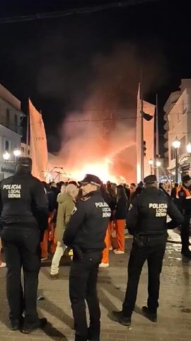 Three police officers wearing black jackets with the inscription "POLICIA LOCAL DENIA" stand facing away from the camera on a cobblestone street at night.  In front of them, a crowd of people, many wearing orange jumpsuits, are gathered around a large fire that is burning between two tall, white structures resembling sails. The fire is producing a significant amount of smoke and sparks, illuminating the scene with an orange glow. Buildings line both sides of the street, and street lights provide additional illumination. People are walking and milling around in the background.