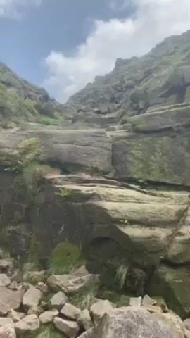 Here is a detailed description of the video:

The video begins with a scenic shot of a rocky, mountainous landscape under a partly cloudy sky. Moss and some sparse greenery are visible on the rocks. The camera then pans down to the ground, where two backpacks are lying among the rocks. 

Next, the video shows the feet of a person walking over a rocky terrain. The person is wearing dark hiking boots and dark-colored pants. The terrain consists of various-sized rocks with patches of grass interspersed. 

The final shot reveals a broad, expansive valley view from a high vantage point. The valley below is covered with lush green fields. The perspective is looking down a steep slope covered with large rocks.