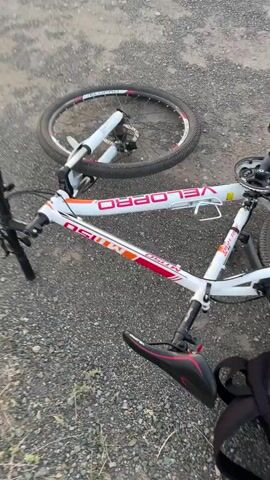 The video begins with a close-up shot of a white bicycle lying on its side on an asphalt road. The bicycle has red and orange markings on its frame and a black seat. The front wheel is slightly detached and lying on the ground. The camera then pans up and to the right to reveal a highway with a yellow line running down the center. The sky is filled with a hazy sunset, with shades of orange and pink. There are some power lines and distant trees visible on the horizon. The camera continues to pan across the highway, showing a few cars in the distance. The video ends with a view of a large metal structure spanning across the highway, possibly a gantry for traffic signs or sensors. The overall scene is one of a deserted highway at dusk, with a fallen bicycle suggesting a recent accident or breakdown.