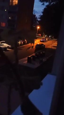A nighttime aerial view shows a street with parked cars and streetlights. A pedestrian walks across the street, followed by another. A car turns the corner.