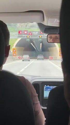 The video shows the interior of a car driving through a tunnel. The view is from the back seat, looking forward through the windshield. The driver, a person with short brown hair and wearing a pink shirt, is visible from the back. In the foreground, the back of two dark car seats can be seen. The tunnel is well-lit with a series of lights along the ceiling. There are two cars ahead of the vehicle, a silver SUV and a dark car. Traffic lights and signs are visible outside the tunnel entrance, indicating a speed limit of 80. The car's navigation system is visible on the dashboard screen. The camera is positioned to capture the forward view and the driver's reflection in the rearview mirror.