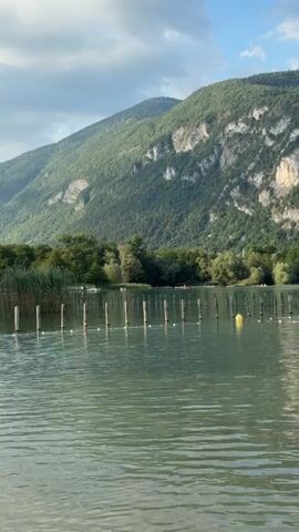 A wide shot captures a tranquil lake with a backdrop of lush green mountains. The water is a calm, muted green, with gentle ripples distorting the reflections of the sky and trees. In the foreground, a line of wooden posts extends into the lake, some topped with white markers. A single yellow buoy bobs in the water to the right of the posts. Further out, a white rope strung with red and white buoys marks a designated area. On the far side of the lake, a dense line of trees forms the transition to the imposing, rocky mountains. The sky is a pale blue with scattered white clouds. In the middle ground, two people are visible swimming in the marked-off area of the lake.
