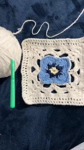 The video shows a close-up overhead view of a white ball of yarn, a green crochet hook, and several crocheted squares with a blue and yellow flower in the center of one. The squares are stacked on top of each other and the video shows hands interacting with the stack of squares. The background is a dark blue fabric.