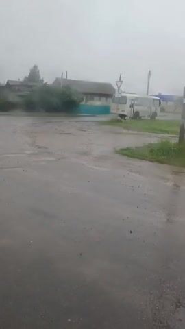 A video taken from a slightly angled, low perspective in an outdoor environment. The ground is wet and appears to be a road or dirt path, reflecting the overcast sky. In the background, there are several structures. To the left, a wooden house with a teal-colored trim is visible, with some greenery and possibly a tree in front of it. Further to the right, a white bus is parked near a building with a blue roof. There are also utility poles with wires present. The sky is uniformly gray and foggy, suggesting poor visibility and possibly rain. The overall atmosphere is damp and dreary.