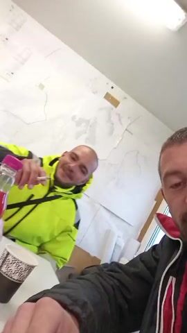 The video opens with a man in a neon yellow jacket sitting at a white table with a cigarette, a bottle of pink juice, and a cup of coffee. The man smiles and raises his fist, and the camera abruptly shifts to the second man, who has a beard and is wearing a black jacket with a red hood. The second man smiles broadly at the camera. The backdrop is a wall with various papers and documents attached. The environment suggests a break room or office space.