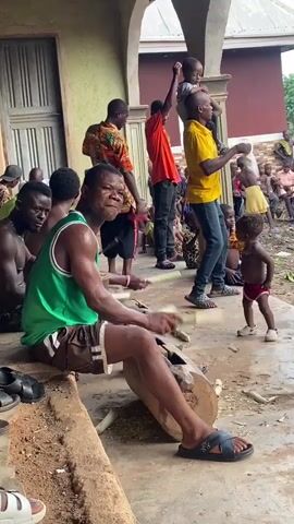 In Ogbu, Nigeria, a group of men and children are gathered outdoors, with several men engaging in a rhythmic performance. In the foreground, a man in a green tank top and brown shorts is seated, intensely drumming on two logs with two wooden sticks. He appears to be sweating from the effort. Behind him, another man in a colorful patterned shirt, and a man in an orange shirt further back, are also holding sticks, seemingly participating in the drumming.

To the right, a man in a yellow t-shirt has a young child perched on his shoulders, holding a white cloth as he sways or dances. A small child in red underwear stands nearby, observing the scene. Other individuals, including children, are visible in the background, some seated, suggesting a community gathering or cultural event. The setting is an outdoor area, possibly a porch or courtyard, with building walls visible. The ground is a mix of dirt and concrete, and the overall atmosphere appears lively and communal under daylight.