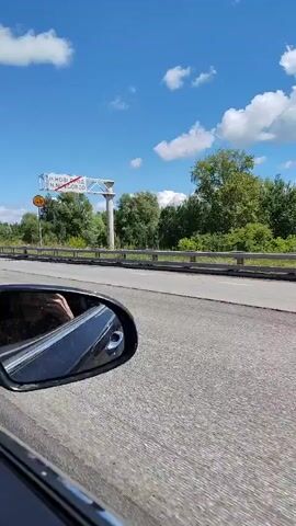The video captures a view from a moving vehicle on a highway under a bright, cloudy sky. The scene is set in Nizhnij Novgorod, Russia, as indicated by a road sign that reads "Н. НОВГОРОД N. NOVGOROD" with a red diagonal line through it, suggesting the city is ahead.

The primary objects in focus are the road, the surrounding vegetation, and other vehicles. A white SUV is prominently featured, driving ahead of the camera's vehicle. The camera's side mirror reflects a portion of the interior and the passing scenery. The weather appears to be fair, with a clear blue sky dotted with fluffy white clouds, suggesting it is daytime, likely late morning or afternoon.

The overall mood is one of travel and transit. There are no visible people, but the presence of vehicles implies human activity. The setting is a typical highway environment with trees and foliage lining the road. A speed limit sign of "40" is visible on a pole, though it is unclear if this applies to the camera's vehicle or a specific section of the road. The video is a snapshot of a road journey through the Russian landscape.