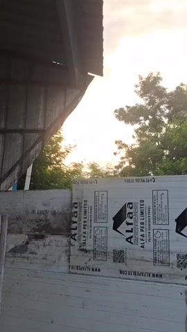 The video shows a partial view of a building's exterior with a large, flat panel leaning against a wall. The panel is white with black text and logos, one of which appears to be "Alfa PEB LIMITED." The panel has several lines of text in small print and QR codes. The sky is visible in the background with clouds and sunlight. Trees are also visible in the distance. The left side of the video shows a portion of a dark, angled structure, possibly a roof or awning, obscuring part of the sky and trees. The overall scene suggests an outdoor construction or storage area.