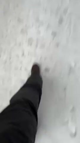 The video shows a first-person view of someone walking on a snow-covered path, with a second person visible further ahead on the path. The person recording the video is wearing dark pants and brown boots. The path is surrounded by snow-covered ground and tall, bare trees. The second person is also wearing dark clothing. The video is taken during daylight.