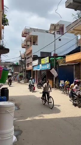 The video shows a street scene in what appears to be a South Asian country, likely India or Nepal, based on the signage and architecture. It is daytime, with a partly cloudy sky. The street is paved with asphalt and shows signs of wear. Buildings line both sides of the street, varying in height and architectural style. Many of the buildings have multiple stories with balconies. Shops with their shutters down or open are visible on the ground floors, displaying goods such as clothing, and possibly food or household items. There are many electrical wires and poles crisscrossing overhead, typical of the region. The street is populated with various modes of transportation. Several people are riding bicycles, including one man in the foreground who is the primary focus of the camera. Others are riding motorcycles or scooters. In the background, a three-wheeled auto-rickshaw is visible, suggesting motorized public transportation. There are also pedestrians walking along the street. The overall impression is of a busy, active urban or semi-urban environment. The camera perspective is at street level, and it appears to be a handheld recording, with some slight movement and focus adjustments.